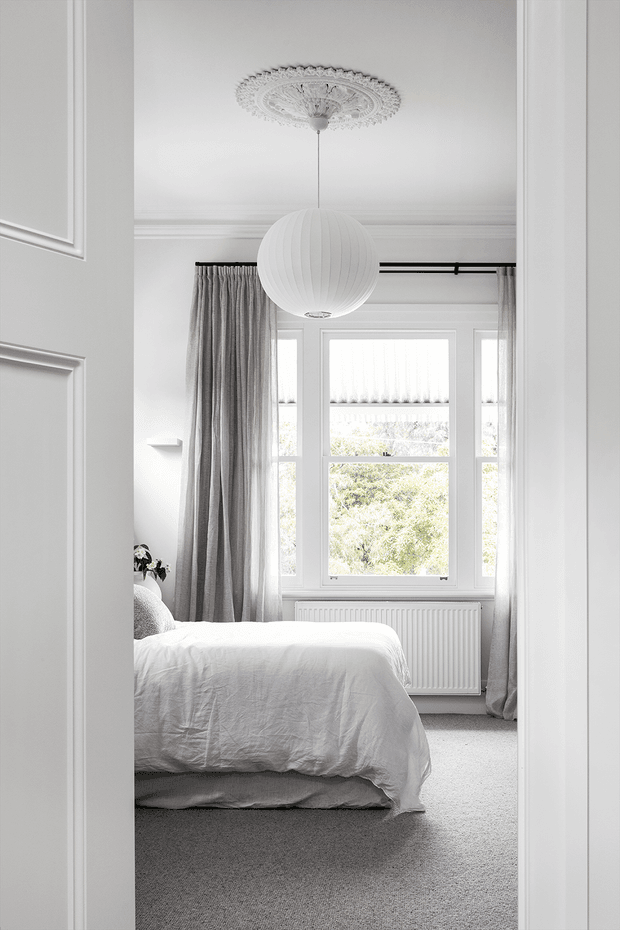 Bright, elegant bedroom viewed through a doorway, featuring a white upholstered or dressed bed, full-height grey curtains, a large multi-pane window, ornate ceiling medallion and cornice detailing, and a spherical pendant light. The room has a soft neutral palette and refined traditional detailing.