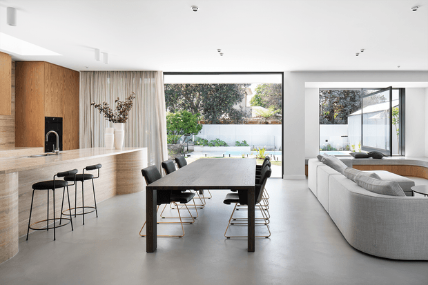 Bright, open-plan contemporary interior combining a kitchen, dining area and living room. The space features a large stone island with black bar stools, timber cabinetry and wall panelling, a dark timber dining table with modern chairs, a white sectional sofa, polished concrete-style flooring, and expansive glazing opening toward a landscaped outdoor area.