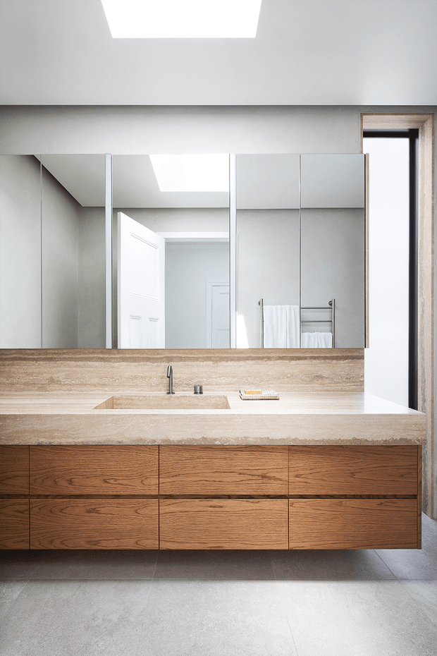 A bright, contemporary bathroom vanity with a floating timber drawer cabinet, pale stone benchtop and integrated basin. Large mirrored cabinetry spans the wall above the vanity, reflecting a doorway and skylight. A towel rail with folded white towels is visible beside a narrow vertical window, while grey floor tiles and a roof skylight contribute to the clean, light-filled design.