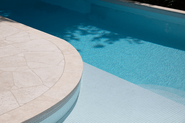 Close-up architectural view of a swimming pool with vivid blue water, pale blue mosaic-tiled interior, and a curved raised circular platform or pool edge finished in light natural stone. Strong geometric curves and tile patterns dominate the composition.