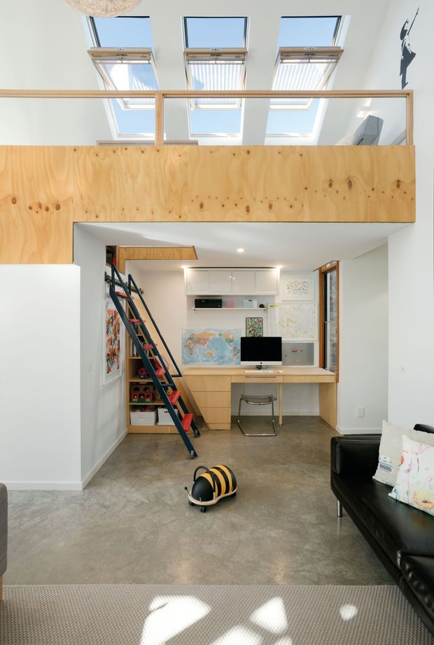 Bright, compact double-height interior showing a children's study and play area beneath a plywood mezzanine. The space has a polished concrete floor, white walls, built-in timber desk and shelving, a dark ladder leading to the upper level, and several skylights providing abundant natural light. A black sofa and children's toys are visible in the foreground.