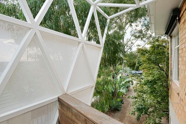 Exterior view from a raised timber deck or balcony beside a brick building, looking through a distinctive white geometric screened structure with triangular bracing and perforated panels. Dense tropical greenery surrounds the property, with a narrow garden path visible below. The image emphasises the architectural screen, balcony edge, and connection to the landscaped exterior.