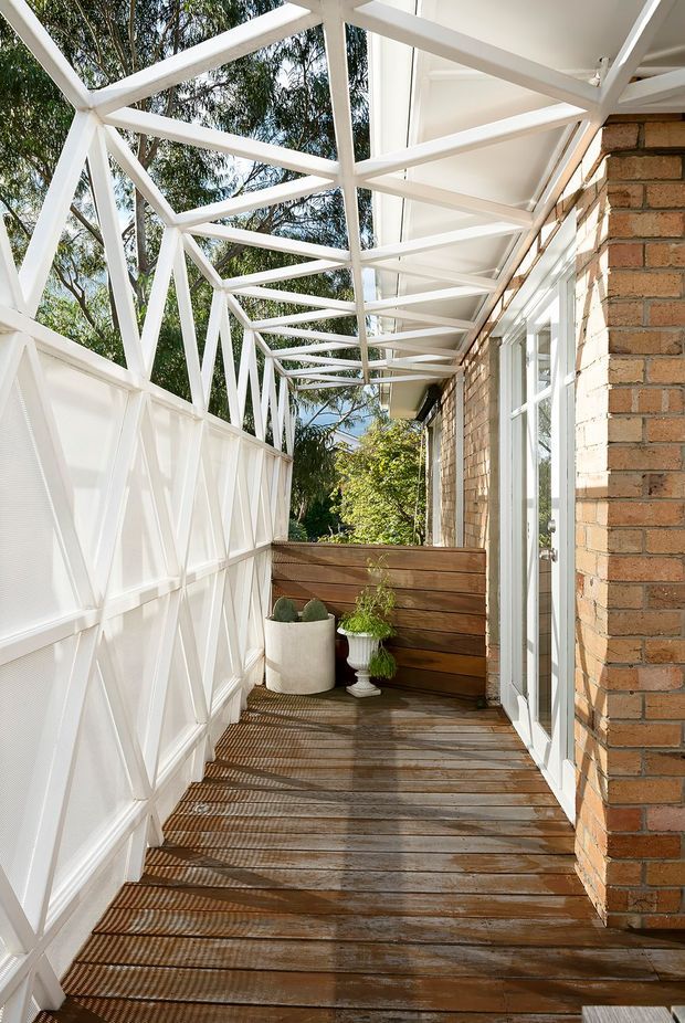 Bright, narrow exterior balcony or terrace walkway beside a brick building. The space has weathered timber decking, a white geometric metal balustrade and overhead trellis-like framing, full-height glazed doors along the brick wall, and a small timber privacy screen with potted plants at the far end. Strong sunlight creates patterned shadows across the deck, with leafy trees visible beyond.