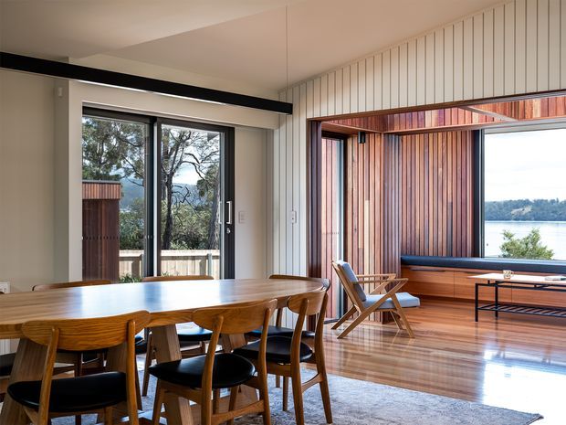 A refined open-plan dining and living area with a long mid-century-inspired timber dining table and chairs in the foreground. The room features polished timber flooring, pale vertically panelled walls, expansive black-framed glazing, and a timber-lined window seat or built-in bench overlooking a body of water and wooded landscape. A low black metal coffee table and lounge chairs occupy the living area, creating a warm, light-filled connection to the outdoors.