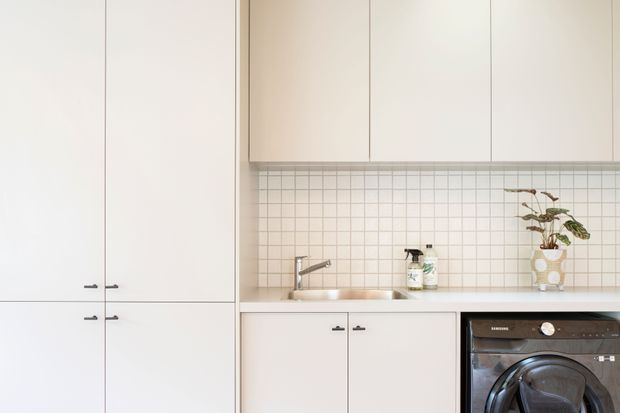 A compact minimalist kitchen or kitchenette with flat-panel off-white cabinetry, integrated tall storage, a small stainless-steel sink and chrome tap, a white square-grid tiled splashback, and a built-in black oven. A small potted plant and cleaning bottles sit on the benchtop.