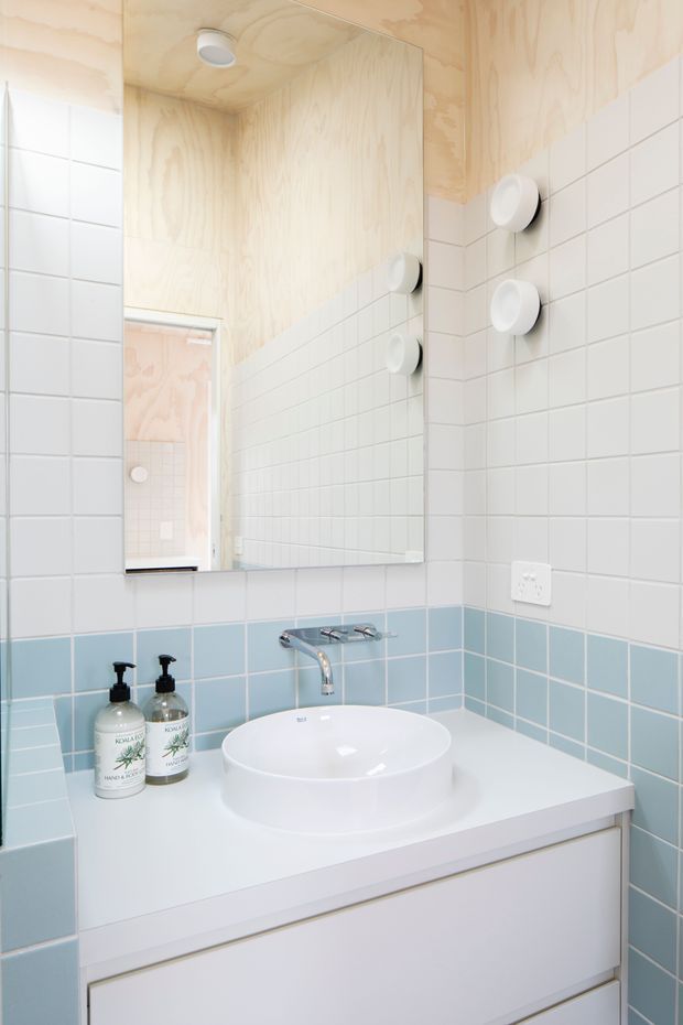 Bright compact bathroom vanity with a white vessel basin, wall-mounted chrome tapware, mirrored cabinet, pale blue and white tiled walls, white built-in storage, and light timber plywood ceiling and wall surfaces. Round white wall lights flank the mirror, creating a clean, simple aesthetic.