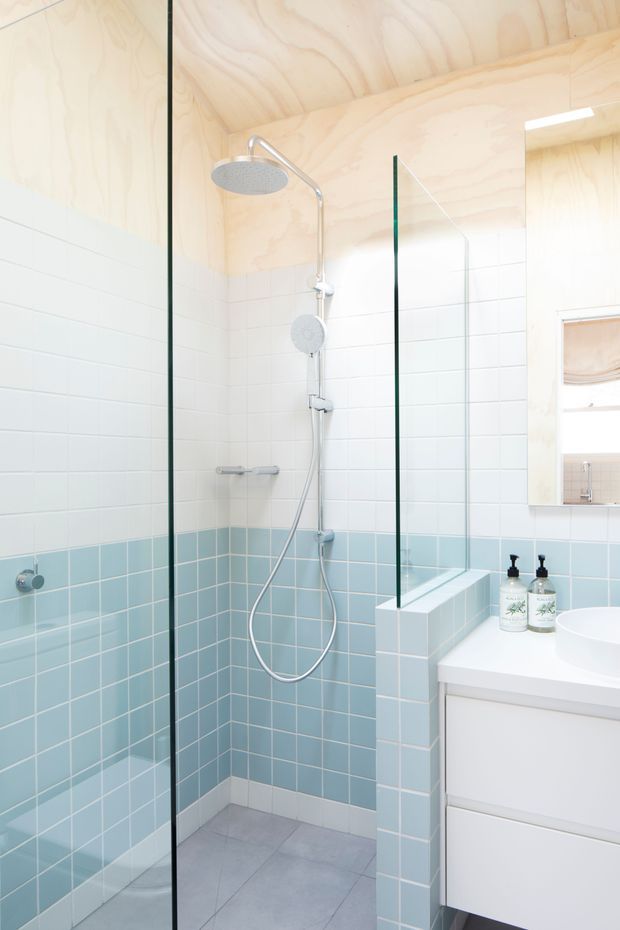 Bright compact bathroom featuring a walk-in shower enclosed by frameless glass, pale blue and white tiled walls, a grey tiled floor, a white floating vanity, and light plywood wall and ceiling lining. The shower has both overhead and handheld fittings, with a small recessed window and mirror visible near the vanity.