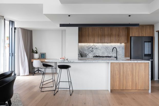 A bright, contemporary open-plan kitchen with a large timber island, white fluted cabinetry, marble-look splashback, integrated dark appliances, and a black linear pendant light. Timber flooring continues into the adjacent dining or living area, with full-height glazing and light curtains providing abundant natural light.