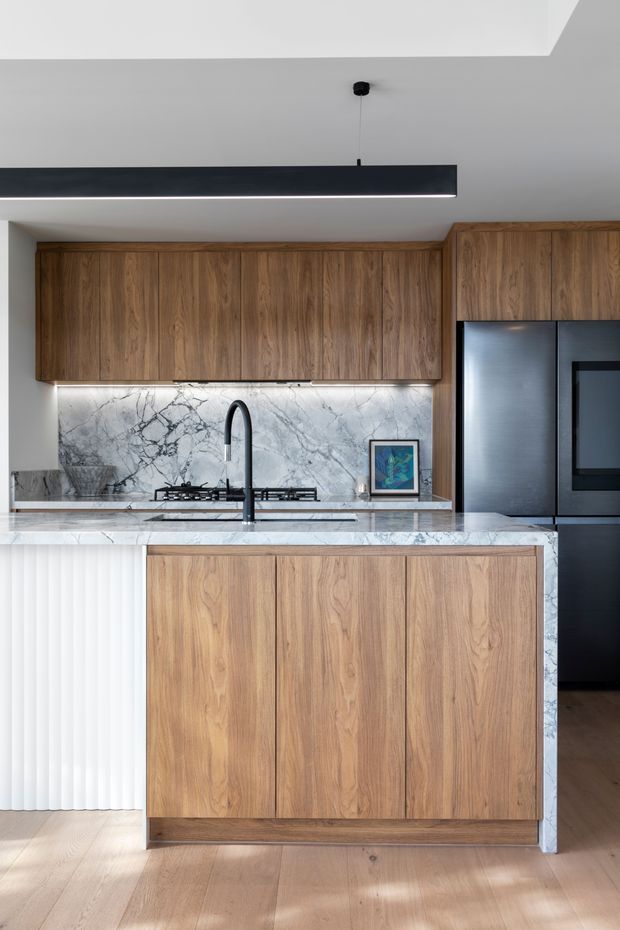 Contemporary kitchen featuring warm natural timber cabinetry, a veined marble-look stone splashback and island benchtop, black appliances and tapware, and a slim linear pendant light. The compact space has light timber flooring, integrated appliances, under-cabinet lighting, and a fluted white panel at the island end.