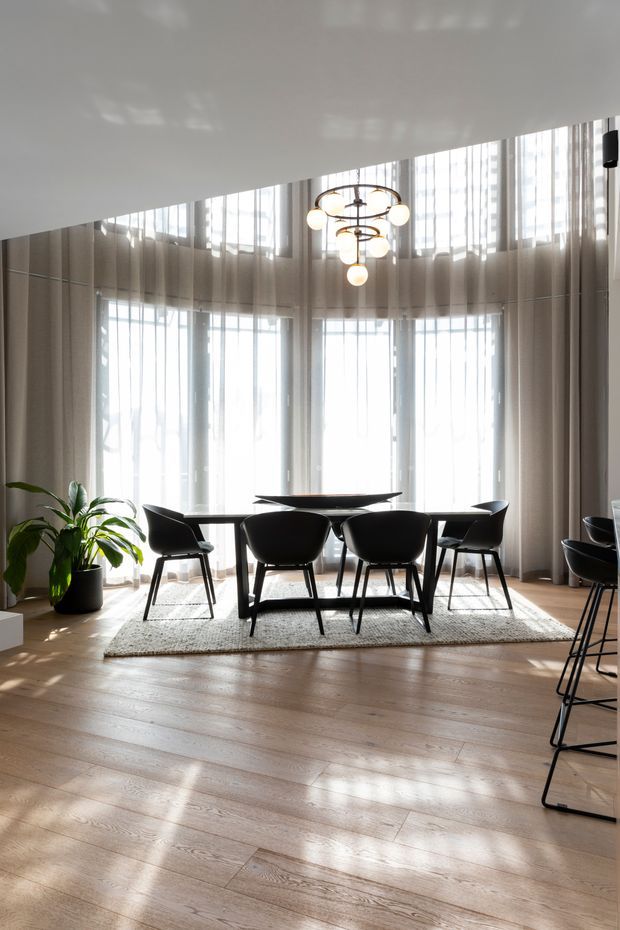 A bright, refined dining area with a long dark dining table and sculptural black chairs arranged on a pale textured rug. Floor-to-ceiling curved glazing with sheer curtains fills the room with daylight, while a multi-tiered spherical pendant light hangs above the table. Pale timber flooring, neutral walls, and a large potted plant complete the contemporary interior.