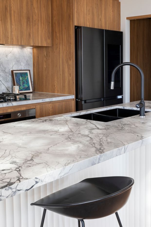 Contemporary kitchen featuring warm walnut-toned timber cabinetry, a large veined white-and-grey stone island benchtop, black appliances and fixtures, an undermount sink with a tall curved tap, and fluted white island-panel detailing. A black bar stool sits at the island.