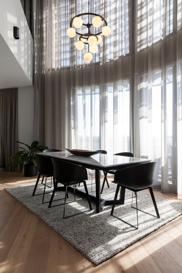 A contemporary dining room with a black rectangular dining table and six black dining chairs arranged on a textured grey rug. Floor-to-ceiling sheer curtains cover expansive glazing, allowing abundant daylight into the double-height space. A sculptural circular pendant with multiple warm globe lights hangs above the table, while a potted plant and neutral drapery add softness. Timber flooring and white walls complete the refined interior.