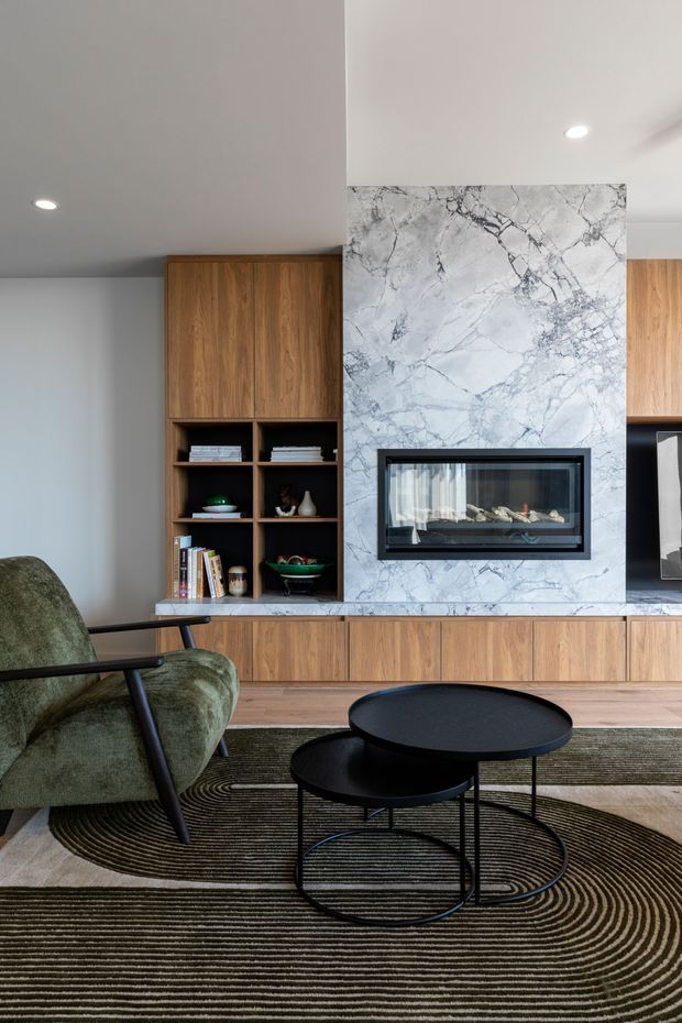 A refined living room featuring a large marble-clad fireplace wall with an inset firebox, integrated warm timber cabinetry and open shelving, a green upholstered armchair, black nesting coffee tables, and a large patterned area rug. Recessed ceiling lighting and a restrained natural palette create a contemporary, sophisticated interior.