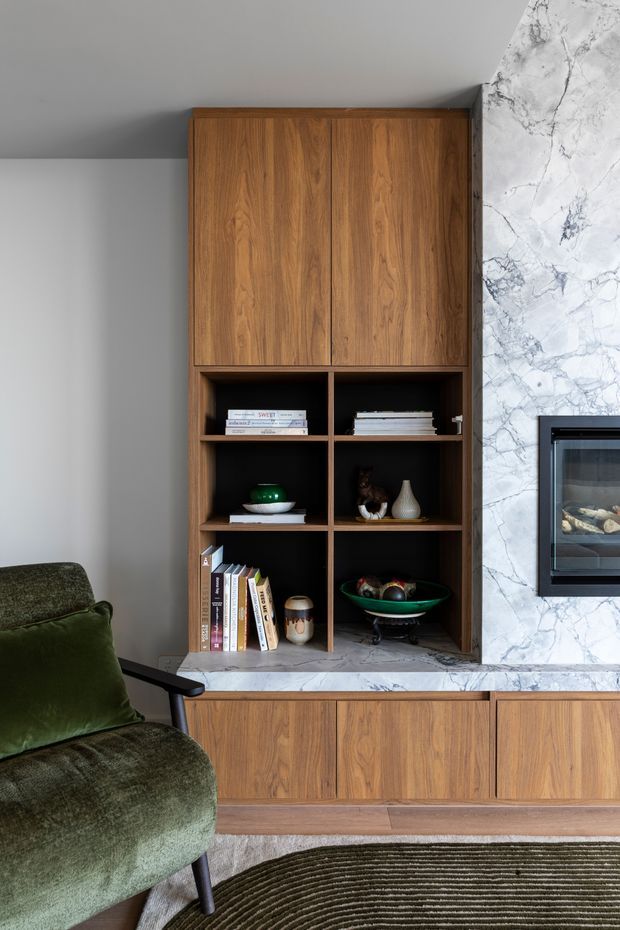 A refined living room joinery wall featuring warm walnut-toned timber cabinetry, open shelving with books and decorative objects, and a pale veined marble surround framing a built-in fireplace. A deep green upholstered armchair and textured round rug add warmth and contrast.