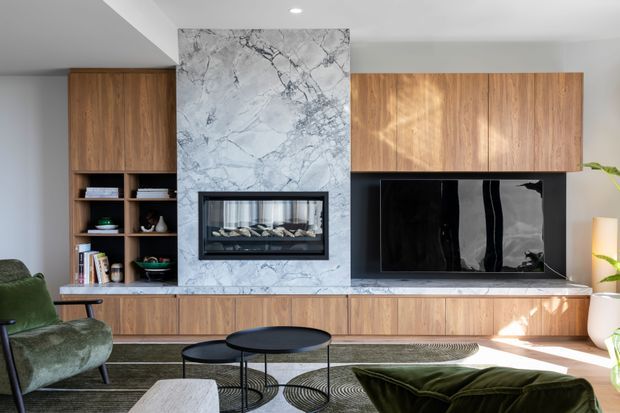 Contemporary living room with a wall-length timber media and storage unit, open shelving, a large black television, and a marble-clad fireplace surround with built-in firebox. Layered coffee tables, a green lounge chair and sofa, and a patterned rug create a warm, refined interior.