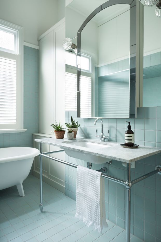 Bright, refined bathroom with pale aqua square wall and floor tiles, a white freestanding bathtub, and a wall-mounted marble or engineered-stone vanity with exposed chrome plumbing and towel rail. A large arched mirrored cabinet dominates the wall above the basin, with white plantation shutters, pale painted cabinetry, and polished chrome fixtures. Small plants and a dark soap dispenser provide restrained accents.