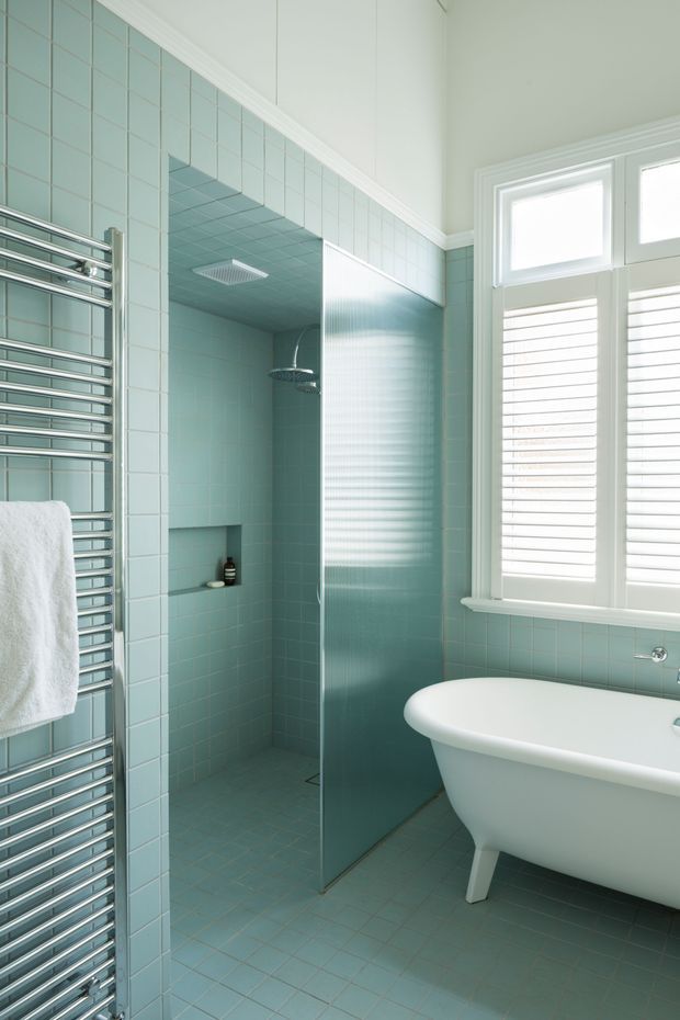 A bright, compact bathroom clad in pale aqua square tiles from floor to ceiling. The room includes a walk-in shower with a rain showerhead and recessed niche, a freestanding white bathtub, a chrome heated towel rail, and a large window with white plantation shutters. White trim and sanitaryware create a clean, soft coastal feel.