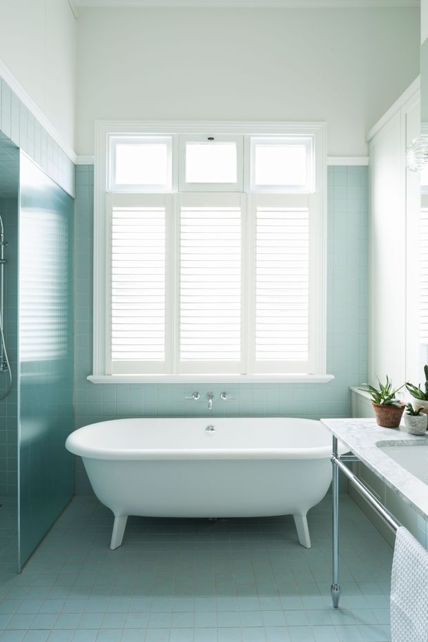 Bright, refined bathroom featuring a freestanding white clawfoot-style bathtub beneath a wide bank of windows with white plantation shutters. Pale aqua square tiles cover the walls and floor, with a glass-sided shower at left and a pale stone vanity with chrome framing at right. The room has white trim, abundant natural light, and a calm, classic coastal feel.