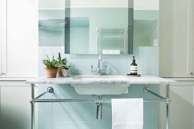 A bright, compact bathroom with pale blue square wall tiles, a white ceramic wall-mounted basin, exposed chrome plumbing, a marble-look stone vanity shelf, and a large mirrored cabinet. A chrome heated towel rail, folded towel, potted plant, and dark soap dispenser add functional and decorative details. The space has a clean, refined, lightly vintage-inspired appearance.