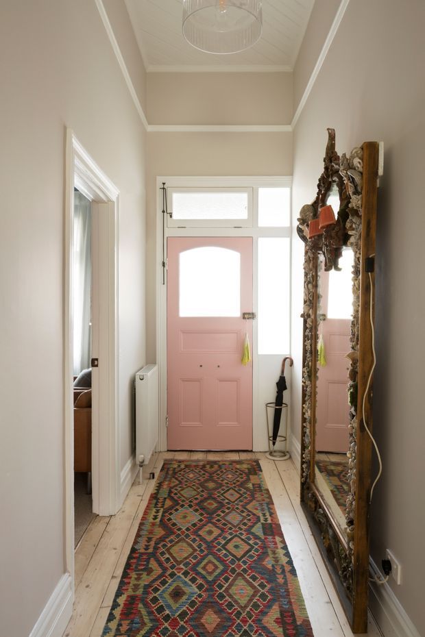 A narrow, light-filled residential entry hall with a pale pink panelled front door, glazed sidelights and transom, white-painted timber floorboards, warm neutral walls, decorative ceiling panelling and a central pendant light. A long patterned runner leads toward the entrance, while a large ornate framed mirror and umbrella stand occupy the right side; an adjoining room is visible through the doorway on the left.