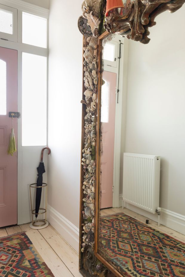 Bright, eclectic entryway with pale timber floorboards, white walls and trim, a muted pink front door with frosted glazing, and a large ornate full-length mirror in a heavily carved frame decorated with shells and natural objects. A patterned geometric runner, umbrella stand, radiator, and reflected doorway complete the space.