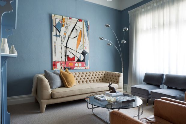 A refined living room with blue painted walls, white-trimmed windows dressed in sheer curtains, and a neutral tufted sofa arranged around a round glass coffee table. Two dark lounge chairs and a brown leather armchair provide contrast, while a sculptural arc floor lamp, built-in shelving, and abstract artwork add decorative character.