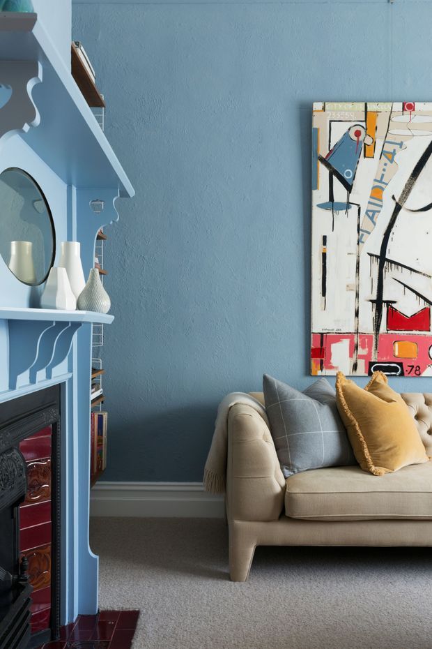 A richly styled living room with a blue-painted textured wall and matching ornate built-in fireplace surround and shelving. A cream upholstered sofa with grey and ochre cushions sits on a neutral carpet, while a bold abstract artwork provides a colourful focal point. Decorative vases, books, and a round mirror add layered character.