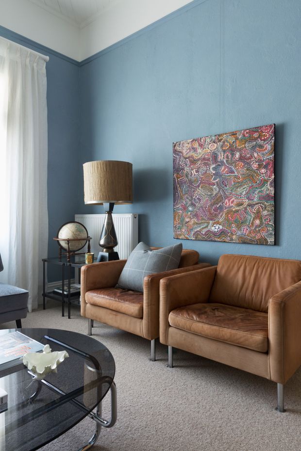 A refined living room with textured blue walls, white sheer curtains, a white painted timber ceiling, and warm brown leather armchairs and sofa. A sculptural table lamp, globe, side tables, colourful patterned wall art, and a glass coffee table complete the layered interior.