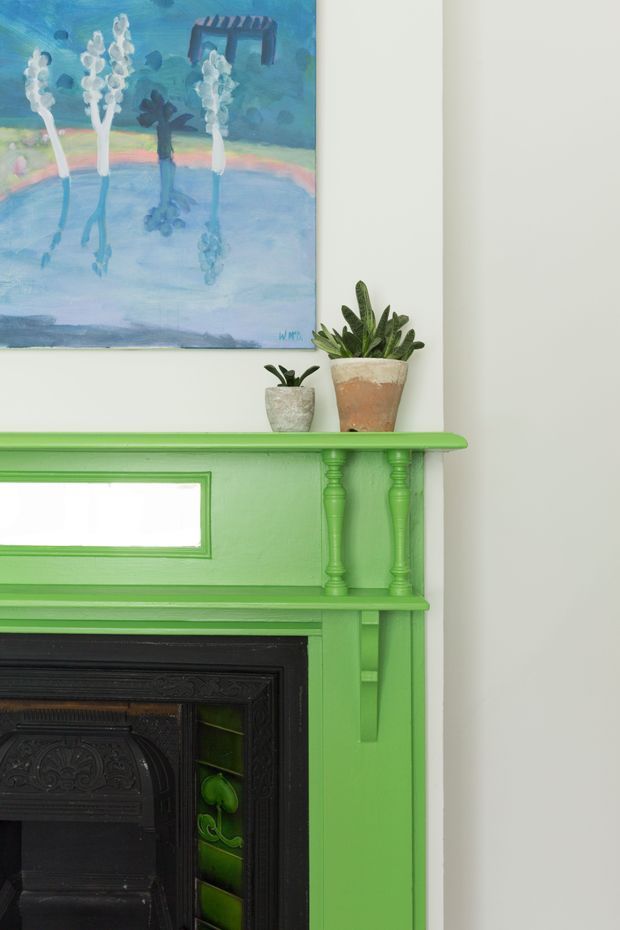 Close-up interior detail of a vividly painted green traditional fireplace mantel with decorative turned supports, moulded trim, and a black ornate fire surround. Small potted plants sit on the mantel, with a colourful abstract botanical artwork above against a white wall.