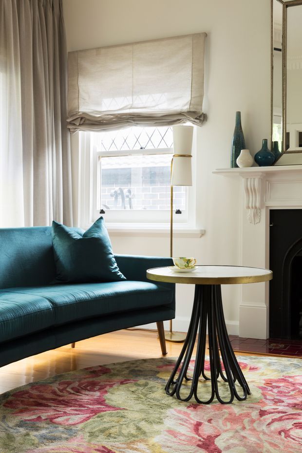 A refined living room with a teal upholstered sofa, patterned area rug, cream Roman blind and full-height curtains, a round brass-topped side table with sculptural black legs, and a white decorative fireplace mantel. A tall mirror and blue glass vessels add detail, while daylight enters through the window.