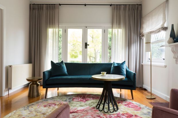 Bright, elegant living room with a deep teal upholstered sofa, patterned floral rug, sculptural round coffee table, and warm timber flooring. Full-height curtains frame glazed French doors, while a bay-style window with a Roman blind and classic wall moulding add refined traditional detailing. The room combines contemporary furniture with classic architectural elements and abundant natural light.