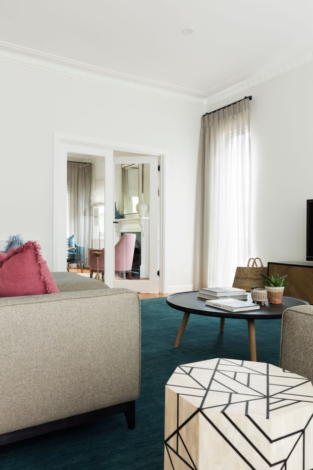 Bright, refined living room with white walls, ornate ceiling cornices, tall sheer-curtained windows, a deep teal carpet, and a neutral upholstered sofa and armchairs. A black oval coffee table, geometric marble-topped side table, books, woven basket, and potted plant create a polished contemporary-classic interior. Mirrored double doors reveal an adjoining room with a fireplace and pink accent chair.