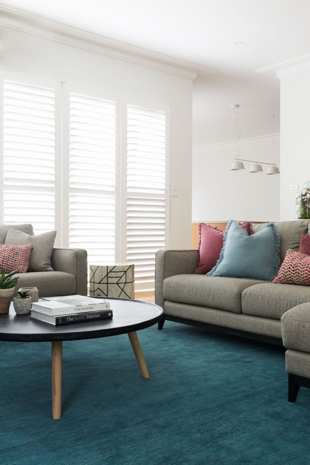 Bright, refined living room with grey upholstered sofas arranged around a round black coffee table. The room features a vivid teal carpet, colourful blue and pink cushions, white plantation shutters, white walls, and decorative ceiling cornices. A minimalist three-light pendant hangs over an adjoining dining area, with a geometric side table and potted plants adding detail.