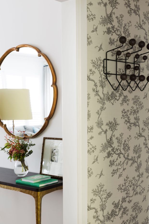 A refined interior vignette featuring a narrow console table with a dark top and brass-toned sculptural legs, an ornate oval wood-framed mirror, a vase of flowers, framed artwork, and stacked books. The adjacent wall is covered in a pale patterned botanical wallpaper with birds, complemented by a striking black geometric wall sculpture. The palette is light and elegant with warm metallic and dark accents.