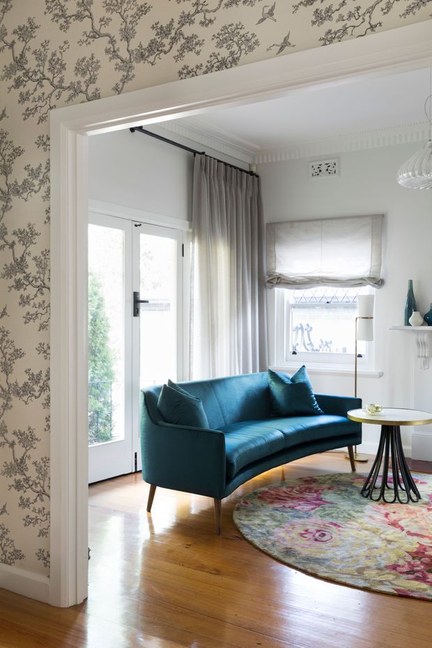 A bright, refined sitting room viewed through a wide cased opening. The room features a deep teal-blue upholstered sofa, polished timber flooring, a colourful round patterned rug, a small round table with a black sculptural base, white walls with decorative cornices and ventilation grille, a glass-panelled exterior door, and a small window with a Roman blind. The adjacent wall and ceiling are covered in a delicate botanical wallpaper, adding a traditional decorative character.