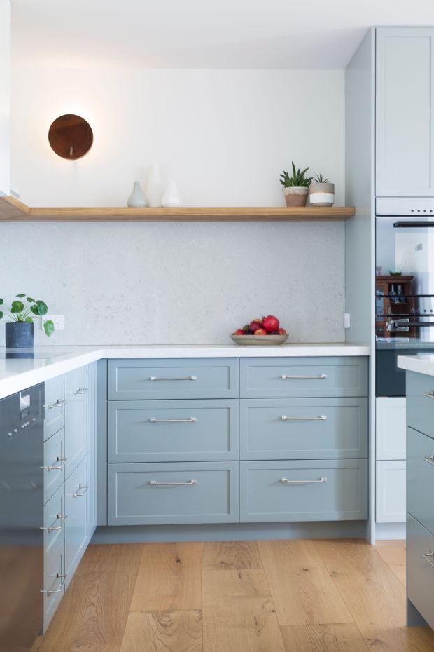 Bright, refined kitchen with pale blue-grey shaker cabinetry, white benchtops, a light textured stone-look splashback, and warm timber flooring. A long open timber shelf displays simple ceramics and potted plants, while integrated ovens and drawers provide built-in storage. The space has a clean, calm, contemporary-traditional feel.