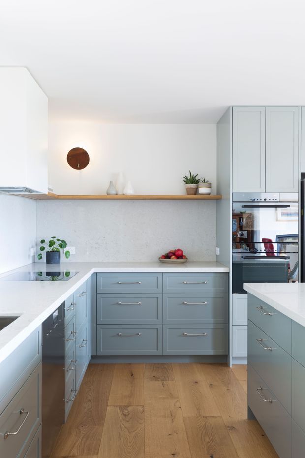 Bright contemporary kitchen with pale blue-grey shaker-style cabinetry, white benchtops, warm timber flooring, and a textured light-grey splashback wall. The layout includes an island and parallel runs of cabinetry, integrated ovens, a cooktop, open timber shelving, plants, and restrained decorative objects.