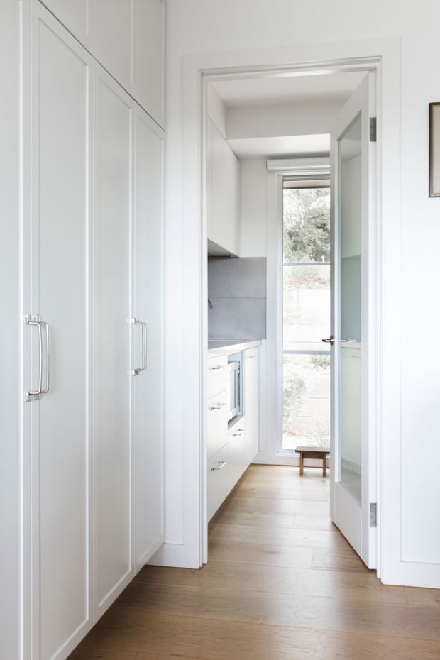 A bright, narrow interior passage leading into a compact kitchen or utility workspace. Floor-to-ceiling white paneled cabinetry with long metal handles lines the left wall, while white cabinetry and a grey backsplash are visible ahead. Pale timber flooring, white walls, and a glazed door or tall window create an airy, clean appearance.