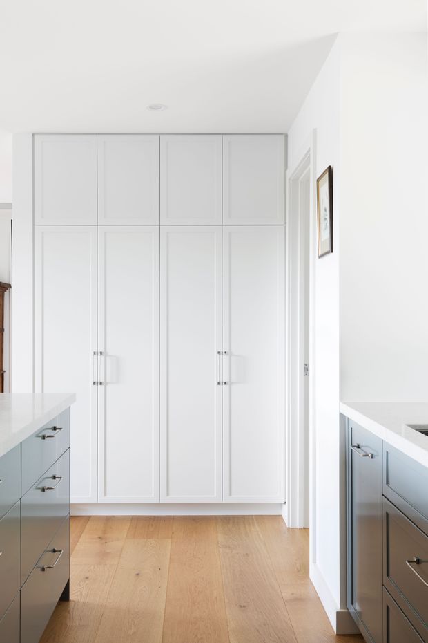 Bright, minimalist kitchen view centred on a full-height wall of white Shaker-style cabinetry. Two pale grey kitchen islands or cabinet runs with light benchtops frame the foreground, while wide-plank light timber flooring and crisp white walls create a clean, airy interior. A doorway and framed artwork are visible on the right.
