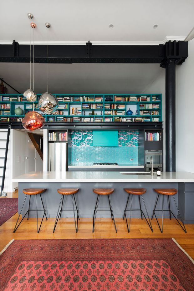A dramatic open-plan kitchen with a long pale island and five sculptural timber-and-metal stools. The room features exposed black-painted structural beams and a large black column, extensive turquoise built-in shelving filled with books and objects, a vivid turquoise patterned splashback, stainless-steel appliances, pendant globe lights, timber flooring, and richly patterned red rugs. A stair ladder and partial adjacent circulation area are visible at the left.