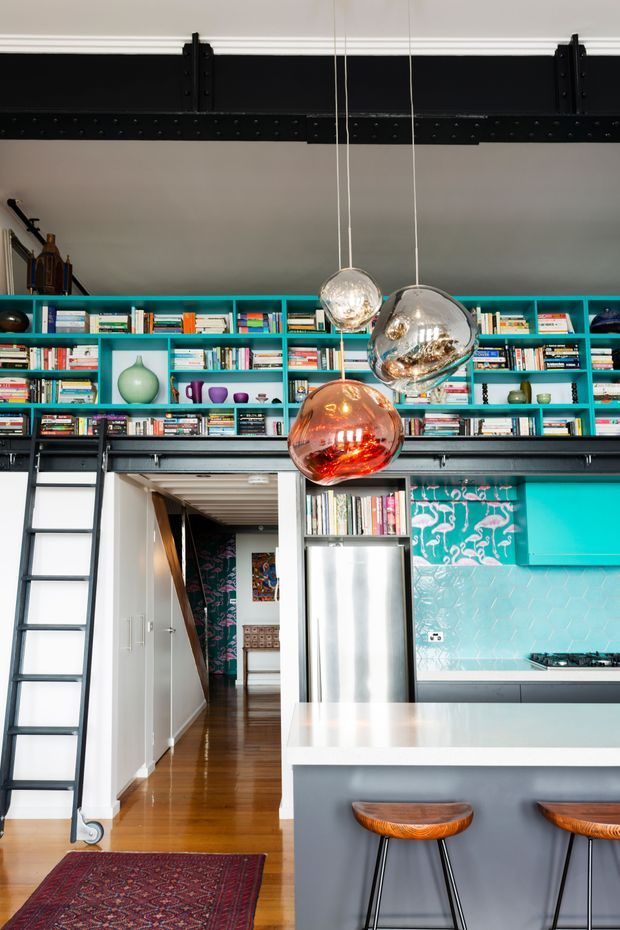 A colourful industrial-style kitchen and living space with a large turquoise built-in bookshelf spanning the upper wall, filled with books and decorative objects. A black exposed steel beam and rolling library ladder run across the room, while three sculptural pendant lights hang over a pale island with wooden stools. The kitchen includes a stainless-steel refrigerator, turquoise cabinetry, patterned backsplash tile, dark benchtops, and polished timber flooring. A hallway and richly patterned wall covering are visible beyond the kitchen.