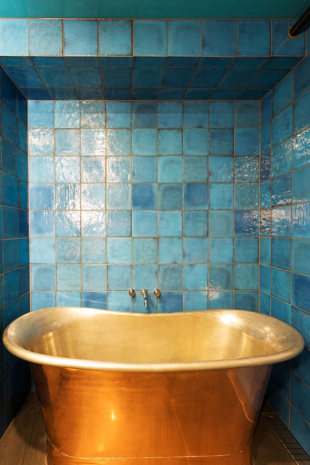 A compact bathroom dominated by a freestanding polished copper bathtub, set against walls and ceiling lined with glossy, irregularly glazed turquoise-blue square tiles. Warm copper fixtures and grout accents contrast with the cool blue tile, creating a richly textured, dramatic bathing space.