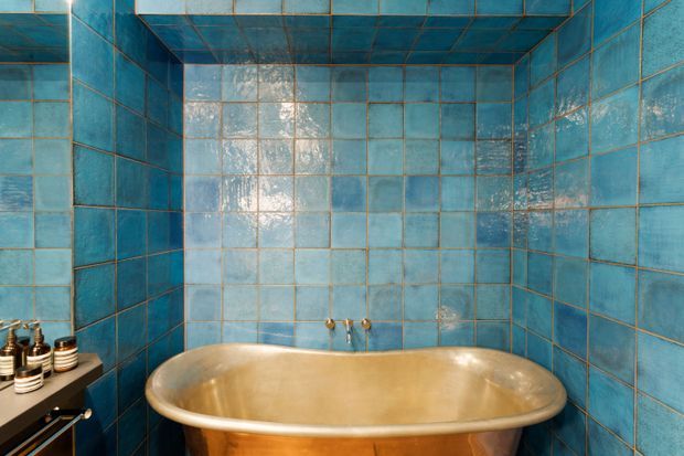 A compact bathroom clad floor-to-ceiling in glossy blue ceramic tiles, including the ceiling, with a freestanding copper-toned bathtub centered against the back wall. A small dark vanity or shelf with toiletries is visible at the left edge. The strong blue-and-copper palette creates a bold, dramatic atmosphere.