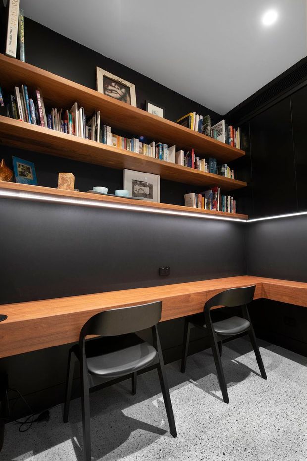 A compact, dark-toned study nook or home office with a long built-in timber desk, two black chairs, open timber bookshelves filled with books, framed artwork, and concealed linear lighting beneath the shelves. Black walls and cabinetry contrast with the warm wood joinery, while the terrazzo-style floor adds a light speckled surface.