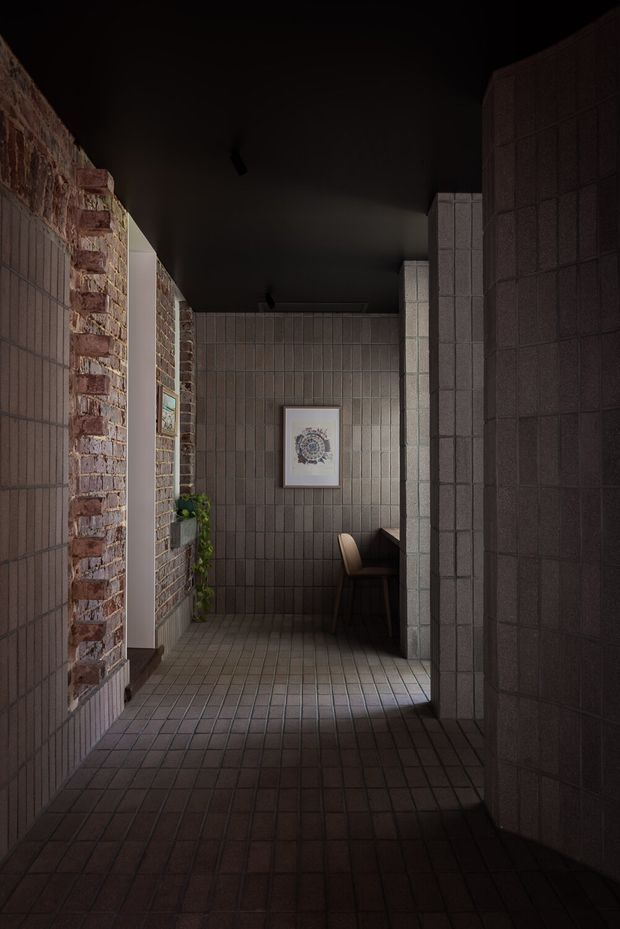 A dimly lit, narrow interior corridor with dark tiled walls and floor, exposed red brickwork on the left, and a black ceiling. The passage opens toward a small built-in seating or dining nook with a chair, dark cabinetry or bench seating, a framed artwork, and a trailing plant. The atmosphere is moody and intimate, with a strong material-focused, industrial character.