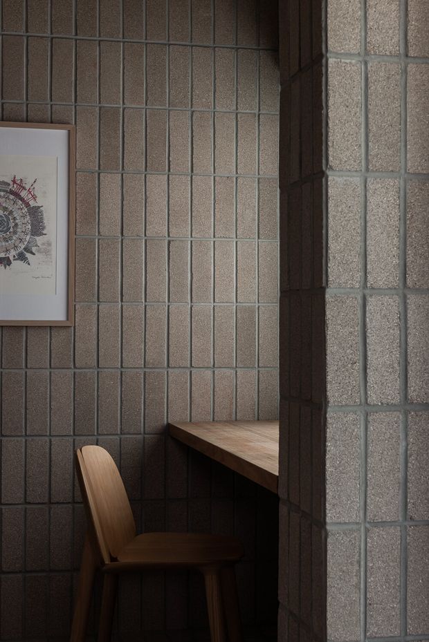 A compact, dark-toned study nook or workspace framed by textured grey masonry tiles. A built-in timber desk projects from the wall beside a simple wooden chair, with framed artwork on the tiled wall. A textured glazed-tile or masonry partition partially screens the nook in the foreground, creating a quiet, enclosed atmosphere.