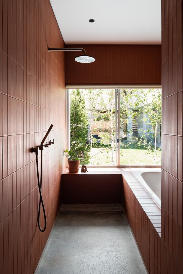 A narrow contemporary bathroom with a walk-in shower and freestanding bathtub. Warm terracotta-brown rectangular tiles cover the walls and built-in bath surround, contrasted with a pale concrete floor and white ceiling. A large window opens the room to a leafy garden, bringing in abundant natural light. Black shower fittings and a small potted plant provide restrained accents.