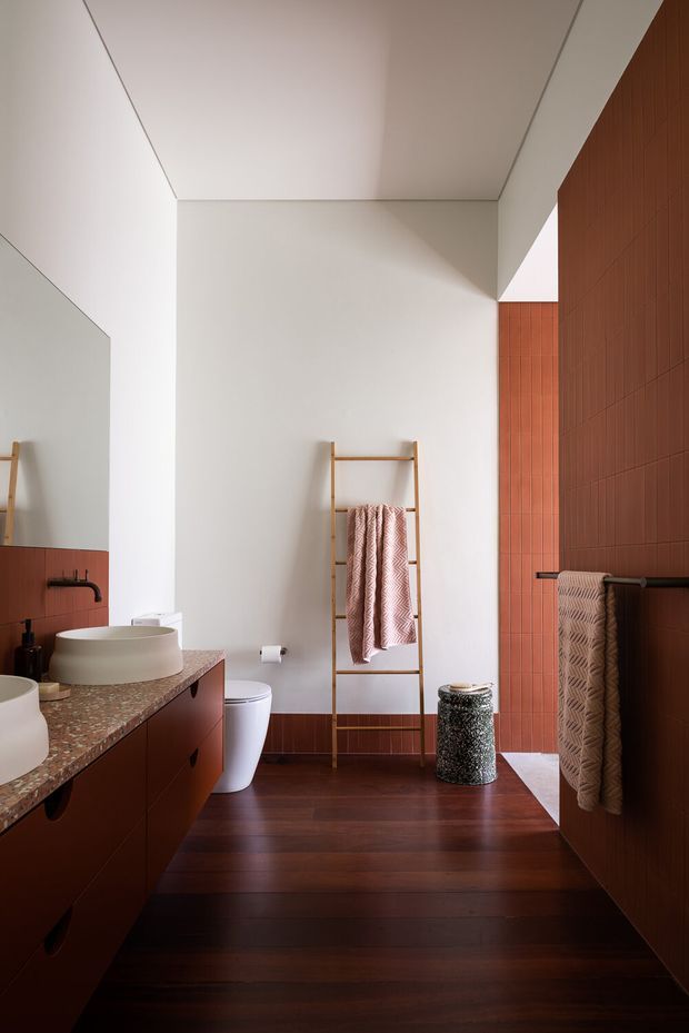 A warm, contemporary bathroom with a dark timber floor, terracotta-red vertical wall tiles, and a matching reddish built-in vanity. The room includes a round vessel basin, black tapware, white toilet, large wall mirror, timber ladder towel rail, and patterned neutral towels. A speckled stone countertop adds texture, while a high white ceiling and strong geometric lines create a clean, refined composition.
