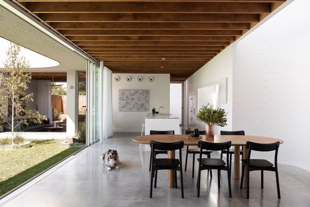 Bright contemporary open-plan dining and living area with a long timber dining table and black chairs, polished concrete flooring, white walls, exposed timber ceiling beams, and full-height sliding glass doors opening to a landscaped courtyard. A compact kitchen is visible at the rear, with strong indoor-outdoor connection and abundant natural light.
