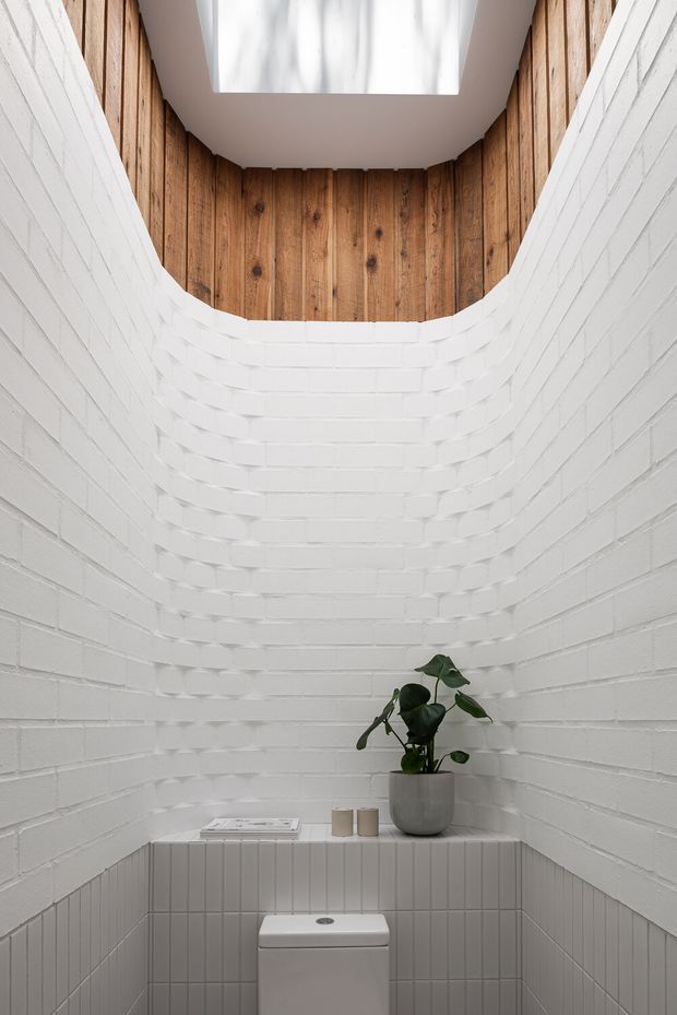 A compact, minimalist bathroom focused on a toilet beneath a skylight. The room has curved white-painted brick walls, vertical white tile wainscoting around the lower section, and a warm timber-lined upper wall beneath the ceiling opening. A small plant and simple accessories sit on the tiled ledge behind the toilet.